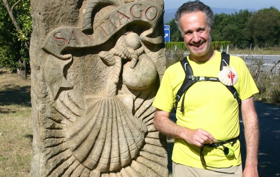 Male hiker in a bright yellow shirt stands smiling in front of stone marker on Spain's Camino de Santiago