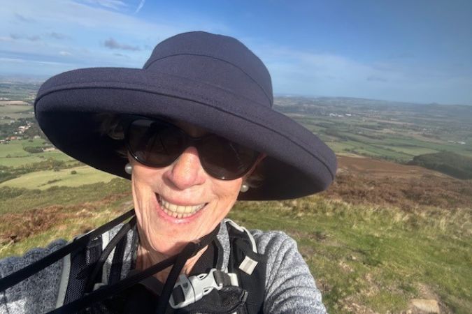 Smiling older woman hiker in a dark blue hat with rolling fells in the distance on the Coast to Coast Path, England