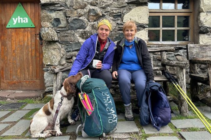 Two hikers sitting outside a stone cottage, with a dog and a backpack, on the Coast to Coast Path in England.