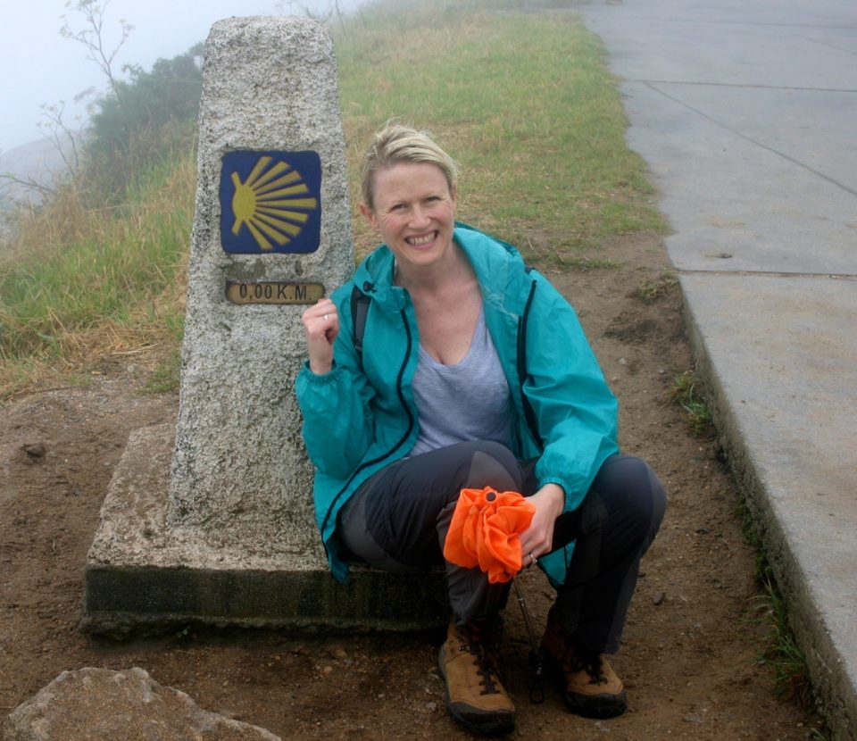 A female hiker sits smiling next to the stone Camino marker at Finisterre - the 'end of the world'.