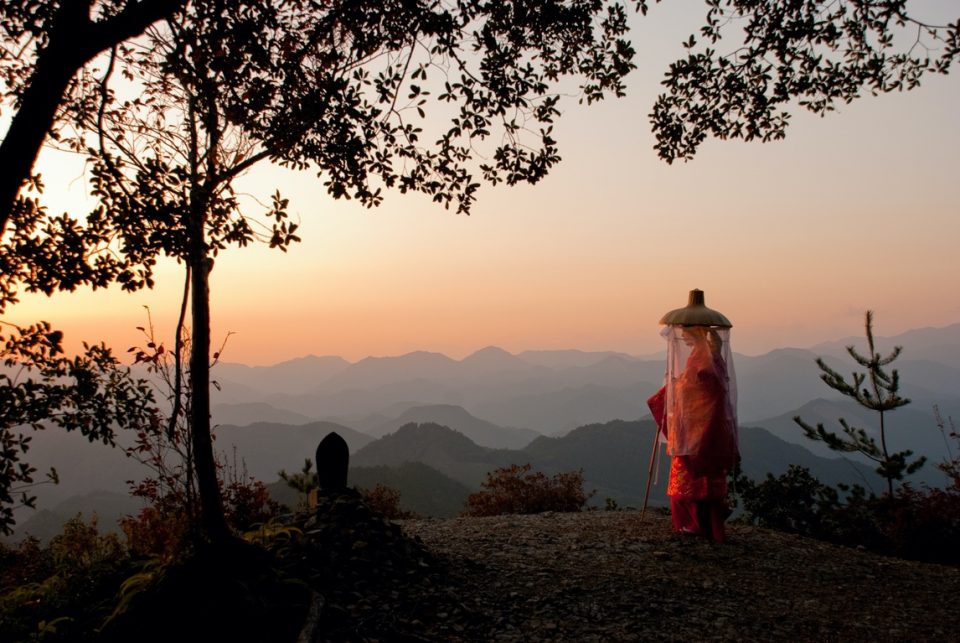 traditional Japanese pilgrim in traditional red dress standing on mountain top looking out over mountain range at sunrise kogumotori hyakkengura pilgrim heian Kii mountains