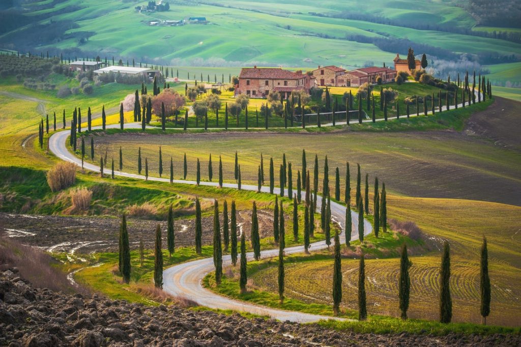 A a path in Tuscany lined with cypress trees leading towards terracotta farm houses and green rolling hills