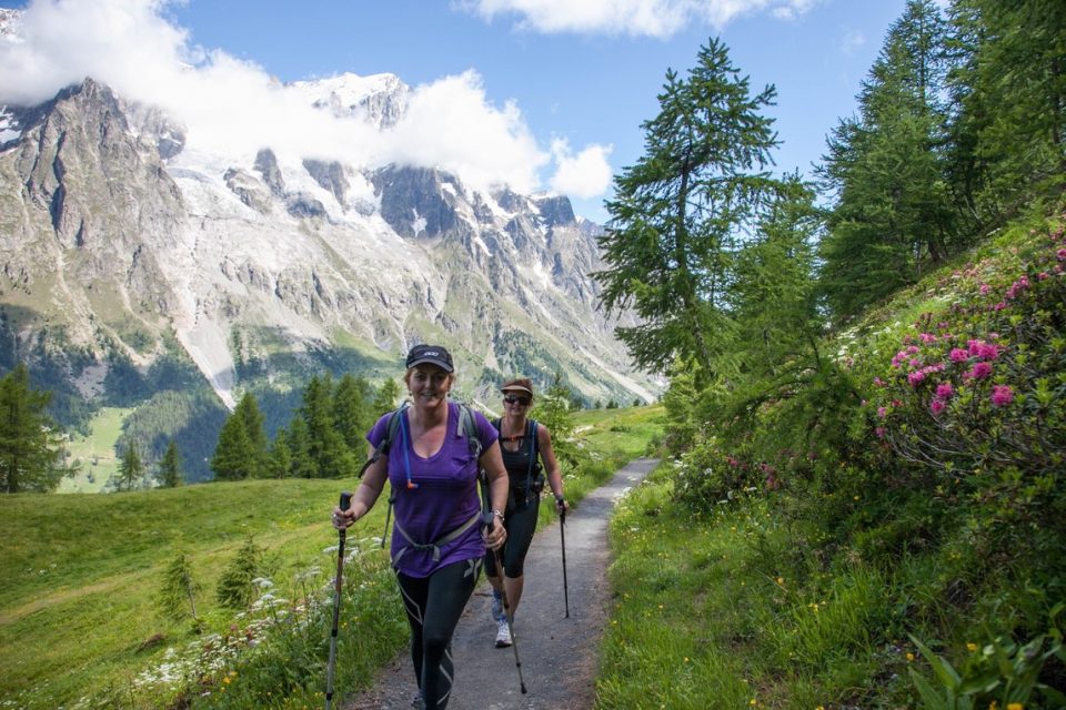 Two female hikers walking towards photographer. They are surrounded by green fields with snow capped mountains in the background.