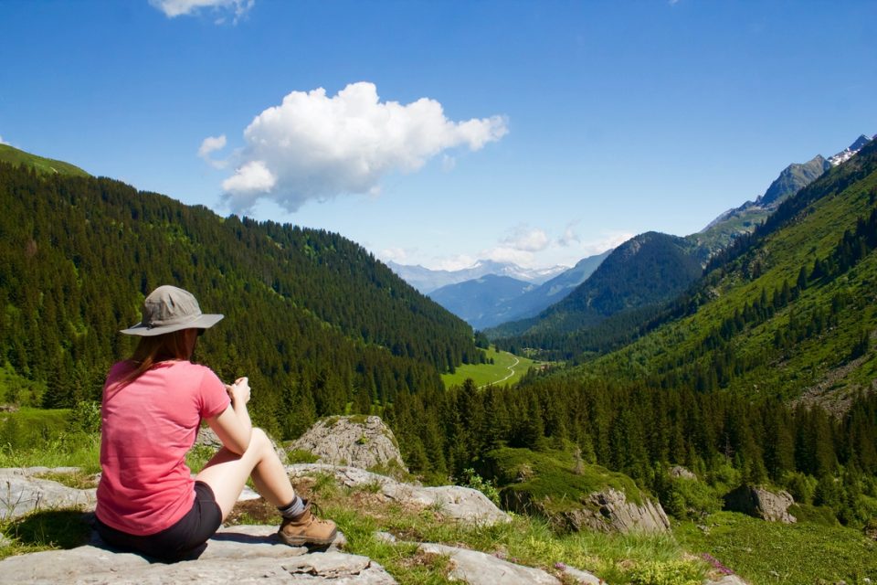 Hiker wearing red tee shirt and hat sitting down looking out over mountains.