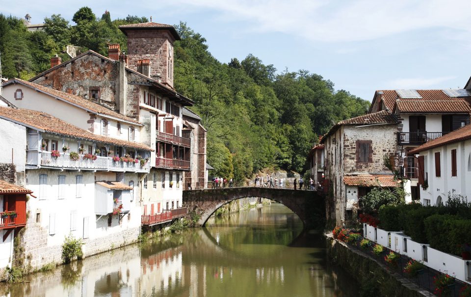 An ancient stone pedestrian bridge in St Jean Pied de Port spans a slow-moving river lined with whitewashed buildings.