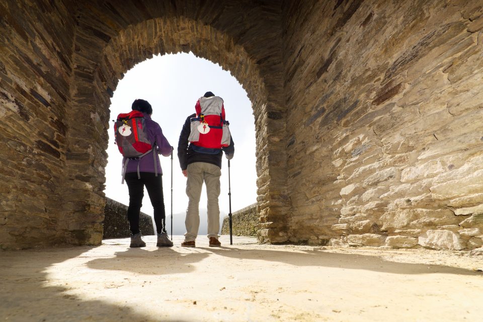 two hikers with their backs to camera, standing in a stone arch way. They have camino shells on their backpacks.