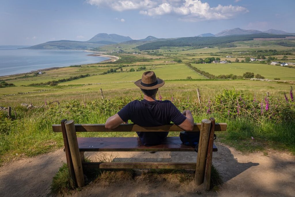 Back of a man sitting on a bench looking at the view below of green rolling hills and mountains on the Isle of Arran, Scotland.