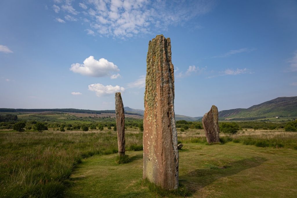 Three large stones called the Machrie Moor Standing Stones on a green expanse with hills in the background on the Isle of Arran, Scotland.