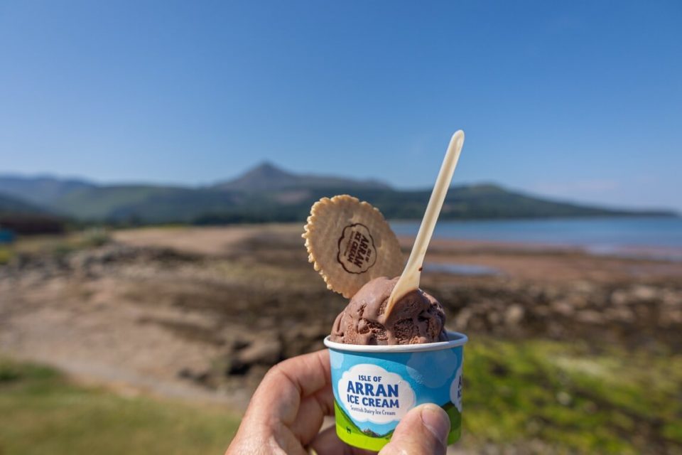 Hand holding small tub of ice cream with a blurry seascape in the background on the Isle of Arran, Scotland.