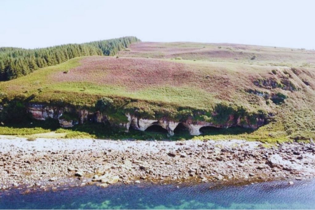 Aerial view of caves dug into a hill with a pebble beach and shore in the foreground.