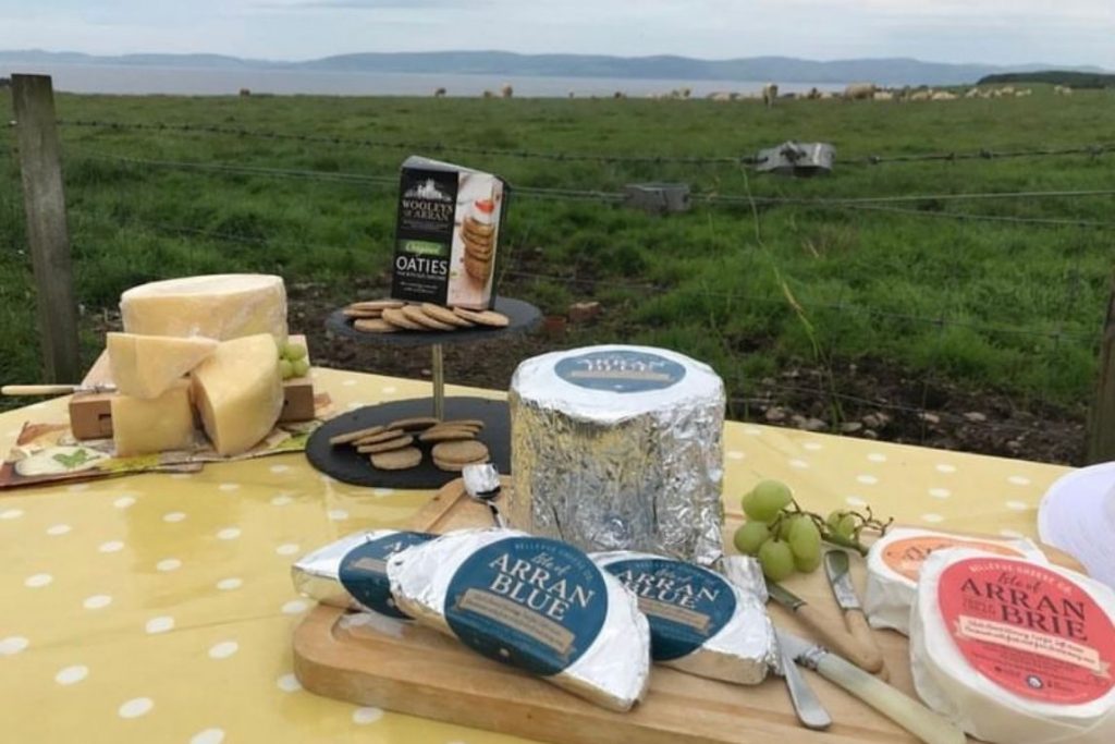 Table with various cheeses and biscuits that is positioned outside in a green field with sheep dotting the background on the Isle of Arran, Scotland.