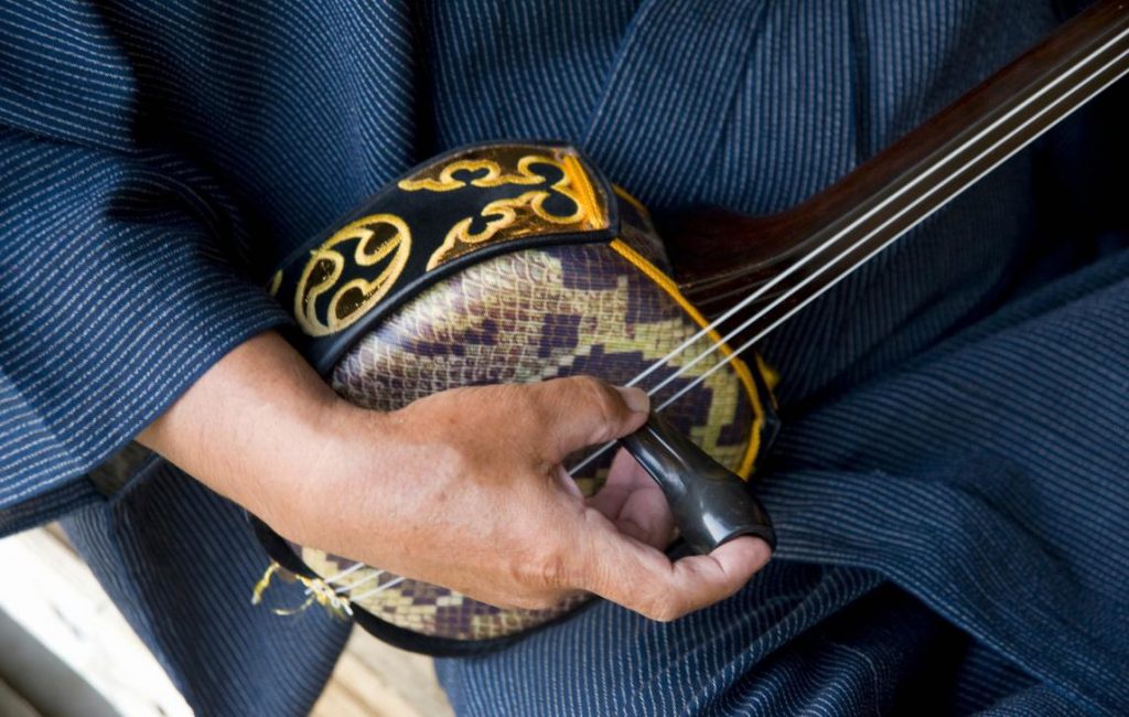 Close up of a man's hand playing a musical instrument that has 3 strings and a snakeskin-covered body; looks similar to a banjo