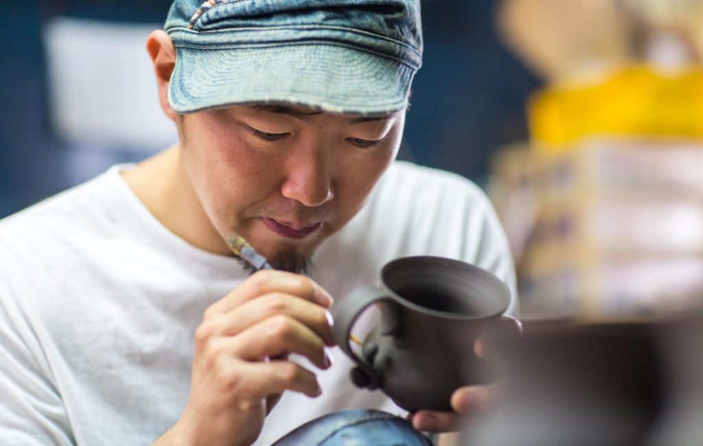 Close up of man wearing a hat putting a finishing image on a cup made from pottery