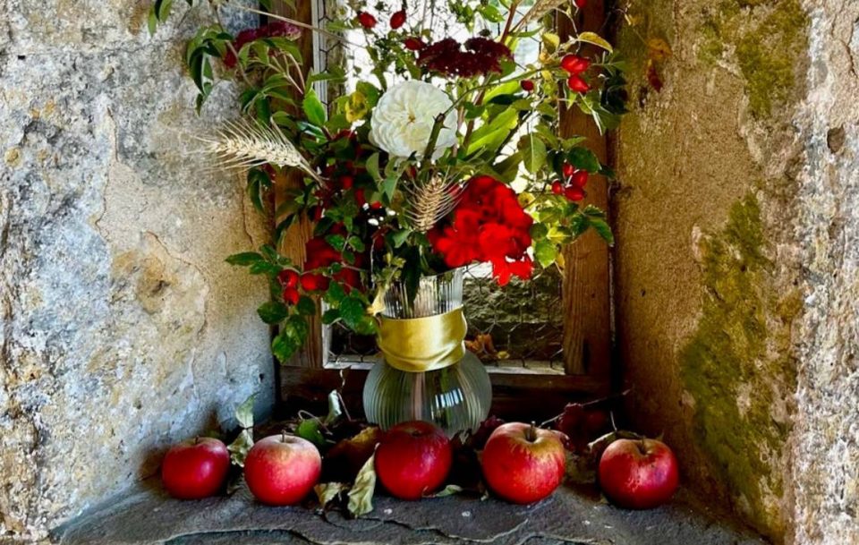 Colorful red flowers and fresh red apples displayed in a vase on a stone ledge.