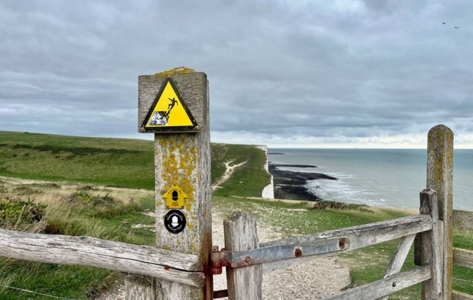 Wooden fence sign by ocean, warning of falling from cliff.