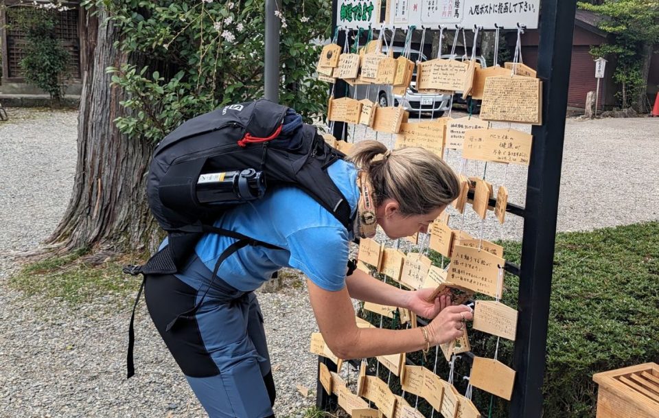A female hiker with a small backpack leans over to read the inscription on a rack of small wooden tiles with Japanese writing on them