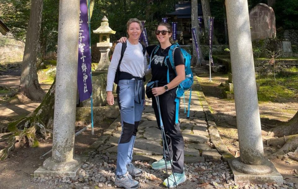 Two female hikers standing in front of a Japanese shrine and smiling at the camera. Kumano Kodo