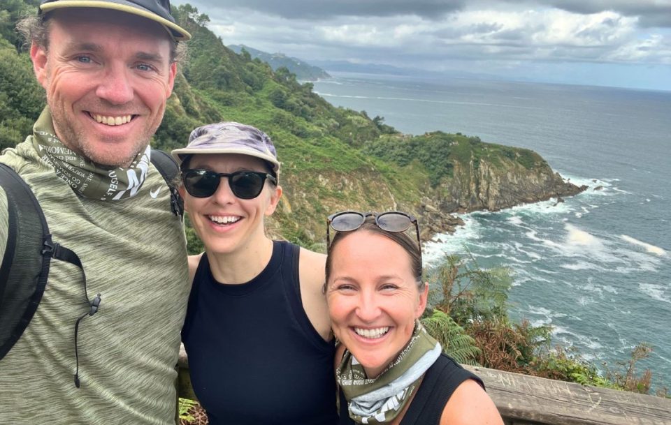 Three individuals smiling and posing for a selfie with a scenic coastal section of the Camino del Norte trail in northern Spain.