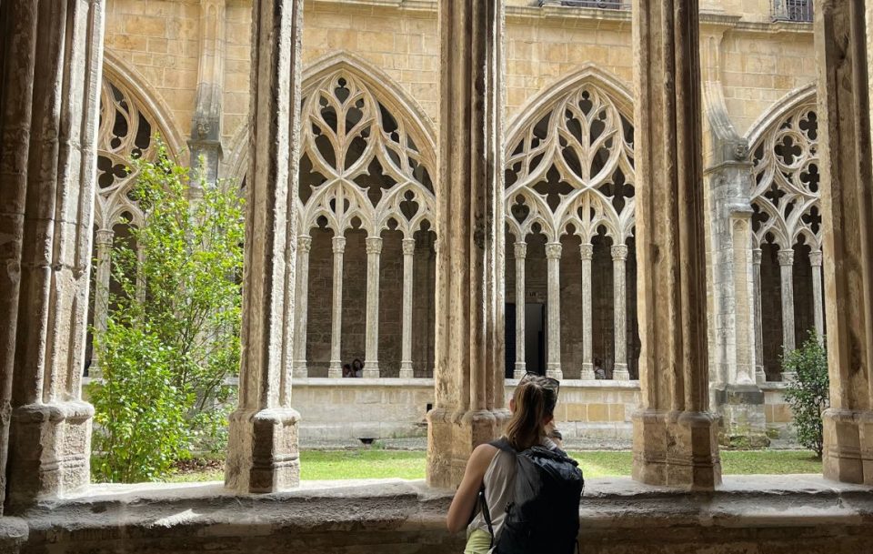 A female hiker with a daypack gazes thoughtfully at at the features of an old church in a courtyard.