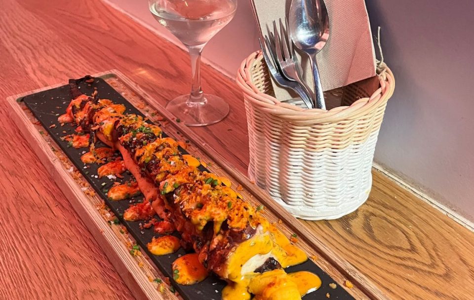 A long piece of meat with a yellow sauce rests on a wooden board, accompanied by cutlery in a basket and a single glass of white wine.