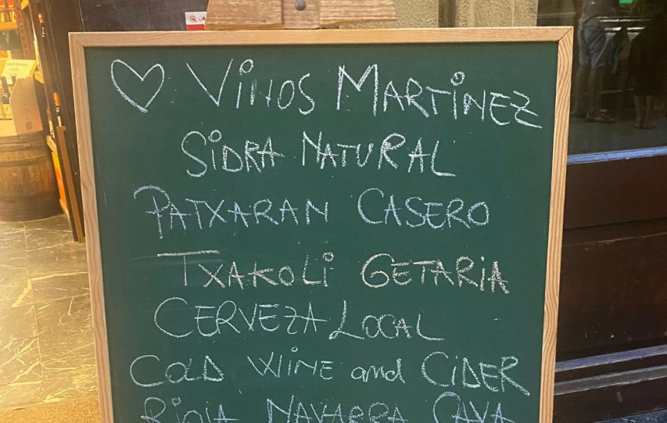 A blackboard sign outside a restaurant displaying menu items including "vinos martinez" and "Sidra natural" in white chalk, set against a dark background.