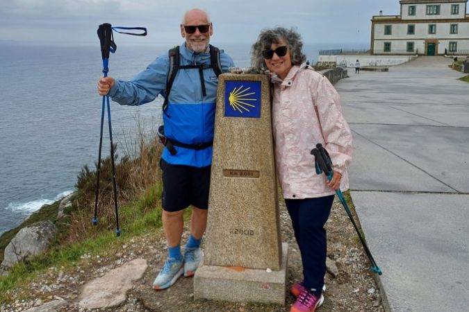Two hikers on the Finisterre coast in Galicia, Spain. They are standing next to a stone marker that has a Camino scallop shell emblem.