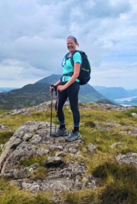Female hiker in blue t-shirt on England's Coat to Coast Path. She is smiling at the camera and there are hills in the background.
