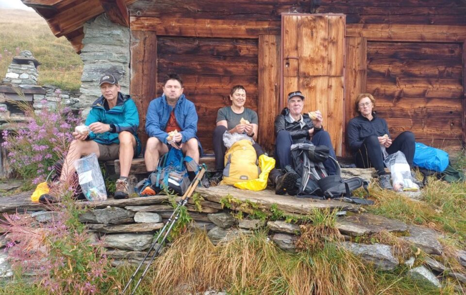 Five hikers sitting outside a wooden cabin having a lunch break along the Walker's Haute Route in Switzerland.