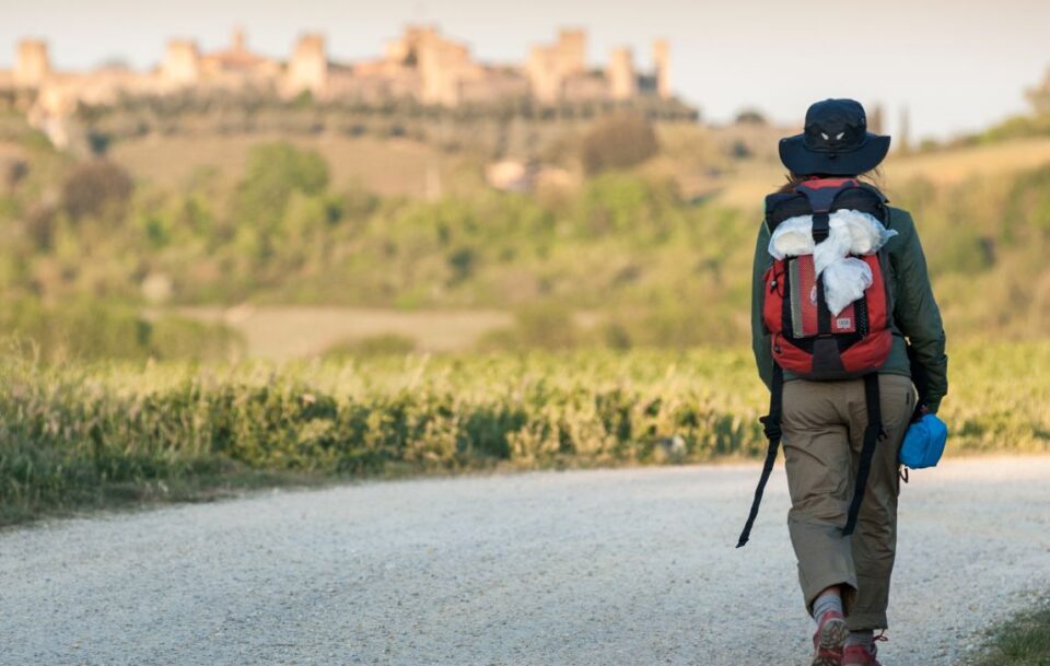 Back view of a female hiker walking on the Via Francigena in Italy with the skyline of a hilltop town in the distance