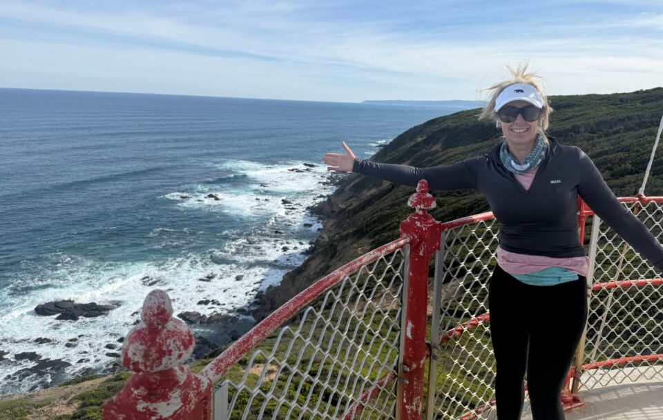 A solo hiker standing on the balcony outside the lantern room at the top of the historic Cape Otway lighthouse, overlooking the ocean.