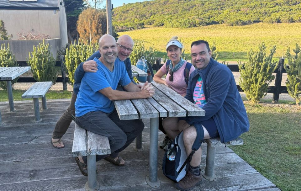Four smiling hikers sitting on a wooden picnic bench with bushland in the background near Castle Cove Lookout on the Great Ocean Walk.