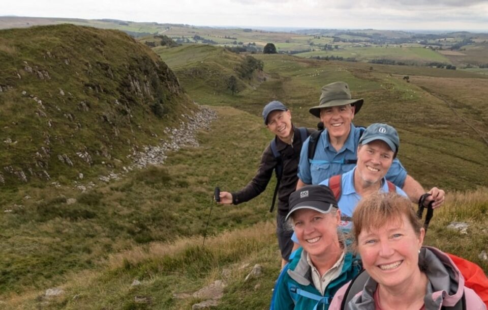A group of 5 hikers smile at the camera in a hilly field dotted with stones on Hadrian's Wall Path