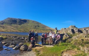 A large group of hikers next to a lake in the Lake District on a sunny day