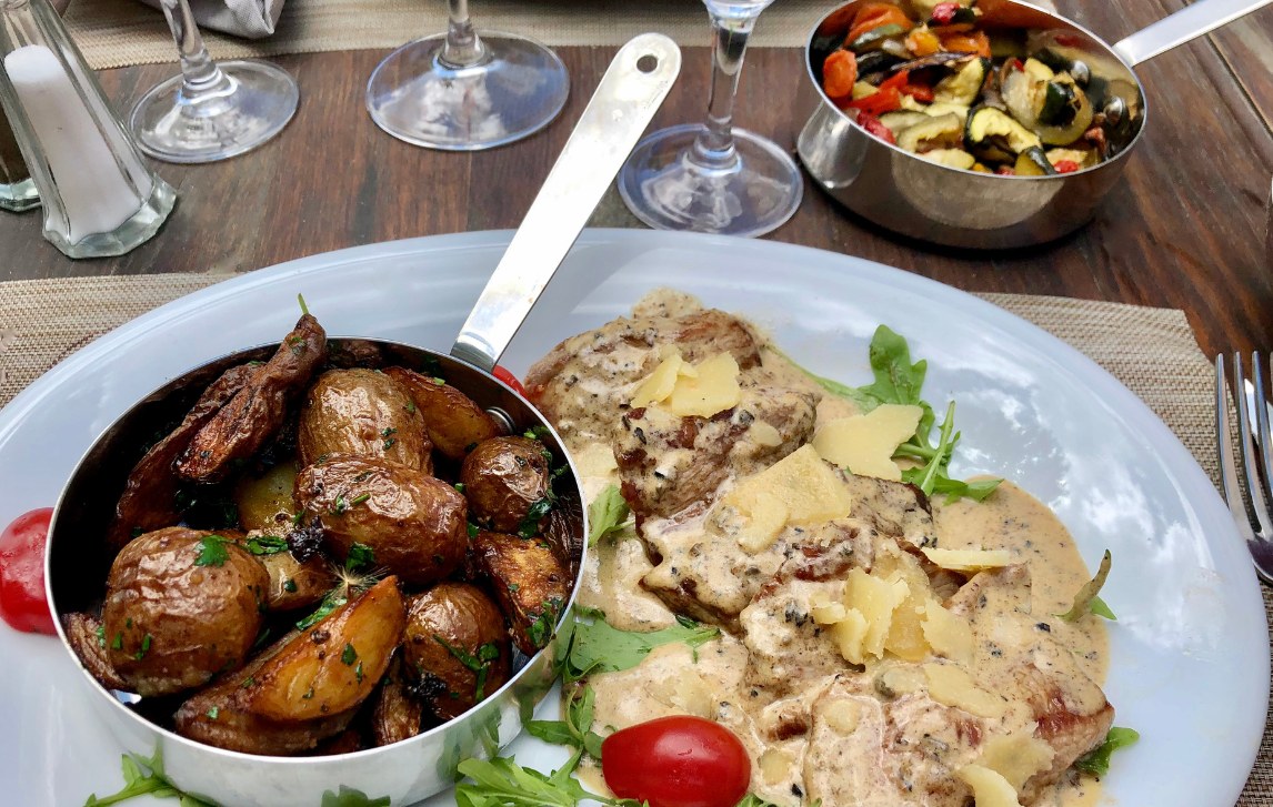 Close-up photo of a plate with veal escalopes cooked in a creamy sauce with truffles and slices of Parmesan cheese, a tomato and rocket leaves garnish, and a small metal pan of potatoes roasted, on a rustic wooden table outside a fine dining restaurant in the south of France.