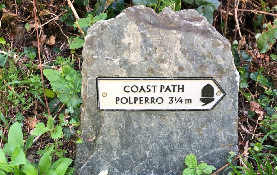 Sign with the National Trail acorn symbol attached to a rock, showing the distance to Polperro on the Cornish Camino path.
