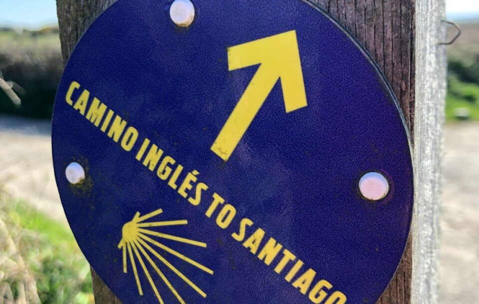 A blue and yellow sign with a scallop shell located on St Michael's Way in Cornwall. It reads: Camino Ingles to Santiago.