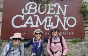 Three female hikers sitting in front of a metal sign that reads 'Buen Camino,' smiling at the camera.