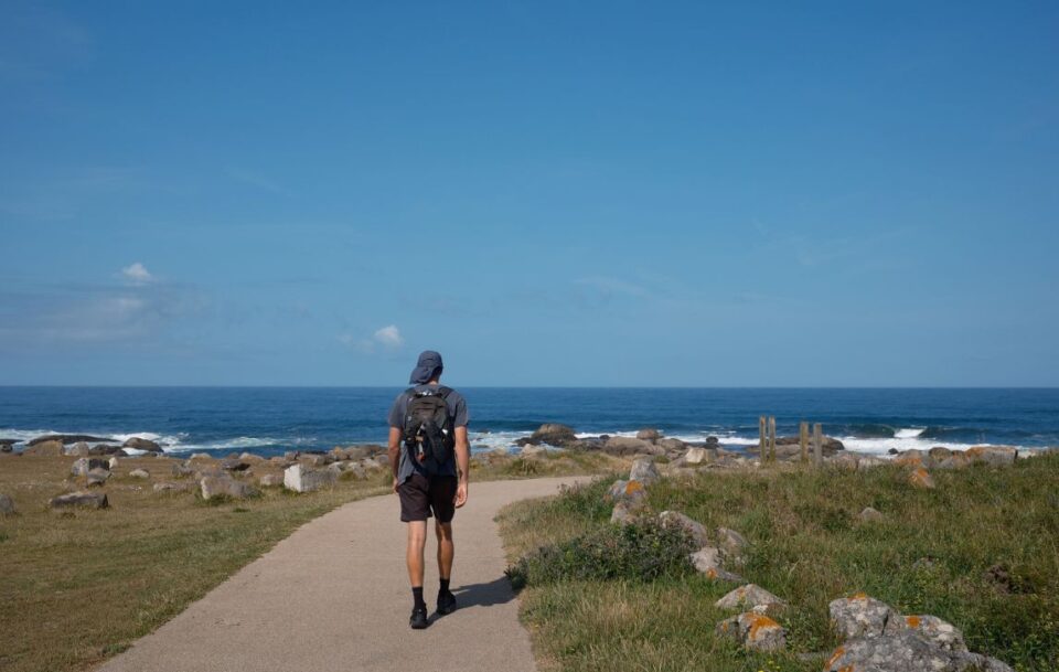 Solo male hiker walking along a paved seashore path of the Portuguese Coastal Camino under a vast blue sky.