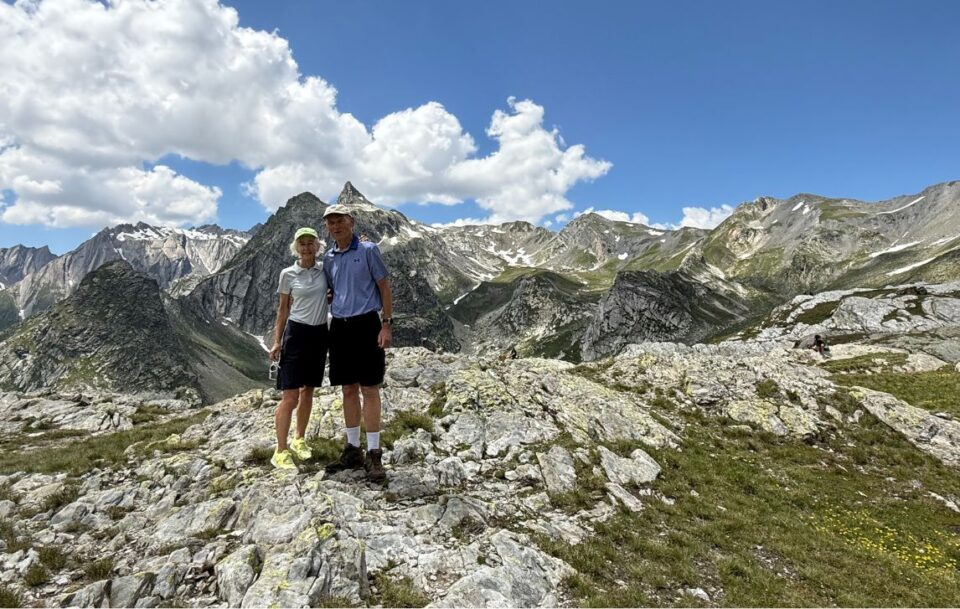 Two hikers standing on the Great St Bernard Pass on the Italian-Swiss border with epic snow-capped mountains behind them.