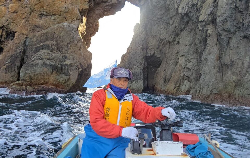 Japanese man steering a small boat along the coastline near the Michinoku Coastal Trail. There are high cliffs and there are white caps on the waves.