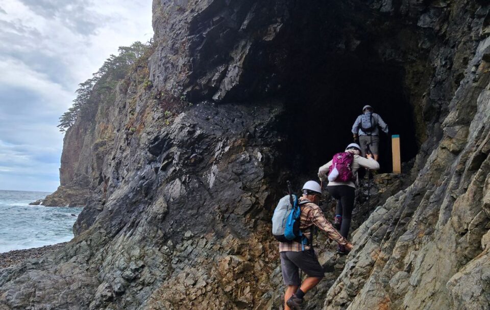 Three hikers entering a dark cave on the shoreline of a beach on the Michinoku Coastal Trail