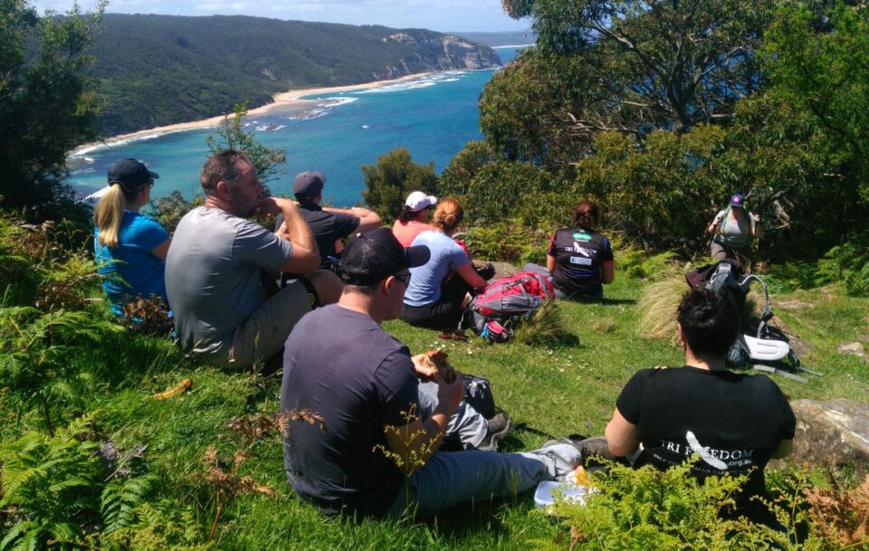 Group of 10 hikers resting on a grassy clifftop on the Great Ocean Walk, taking in sweeping ocean views below.