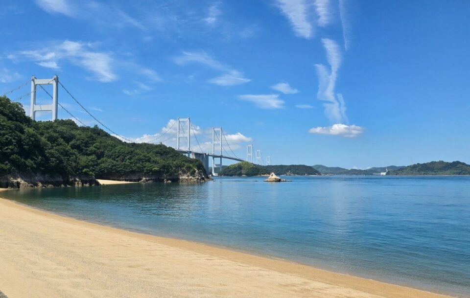 View from the beach of a long suspension bridge between islands along the Shimanami Kaido during daytime