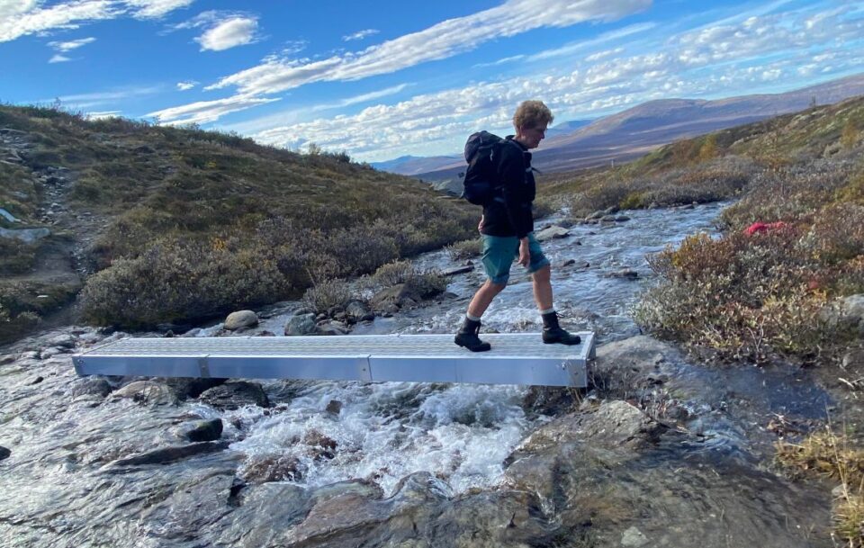 Solo female hiker crossing a fast-running stream on a narrow plank bridge, St Olav's Way Norway