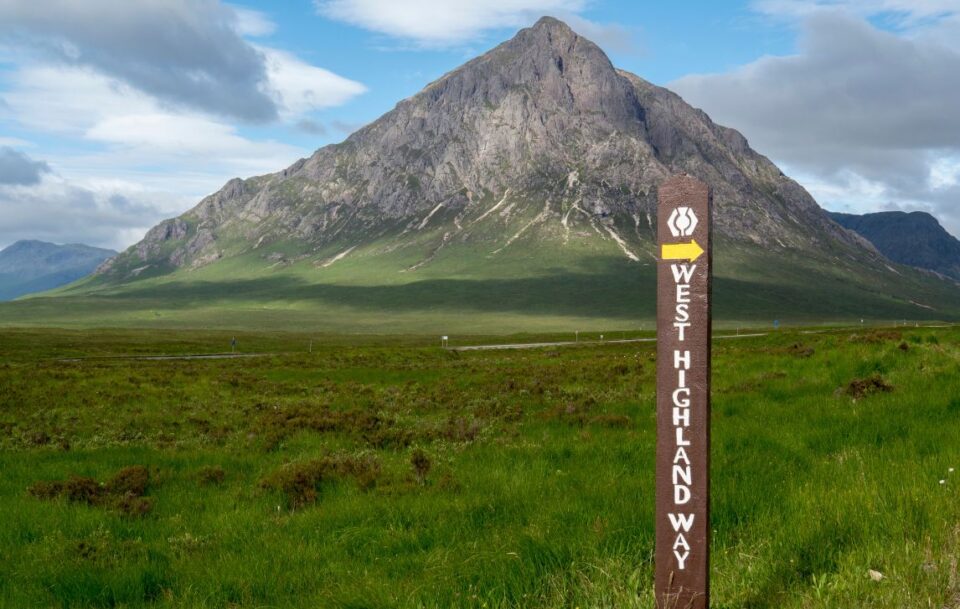 Wooden hiking sign marking the West Highland Way in Scotland, with the scenic mountain peak of Glencoe behind.