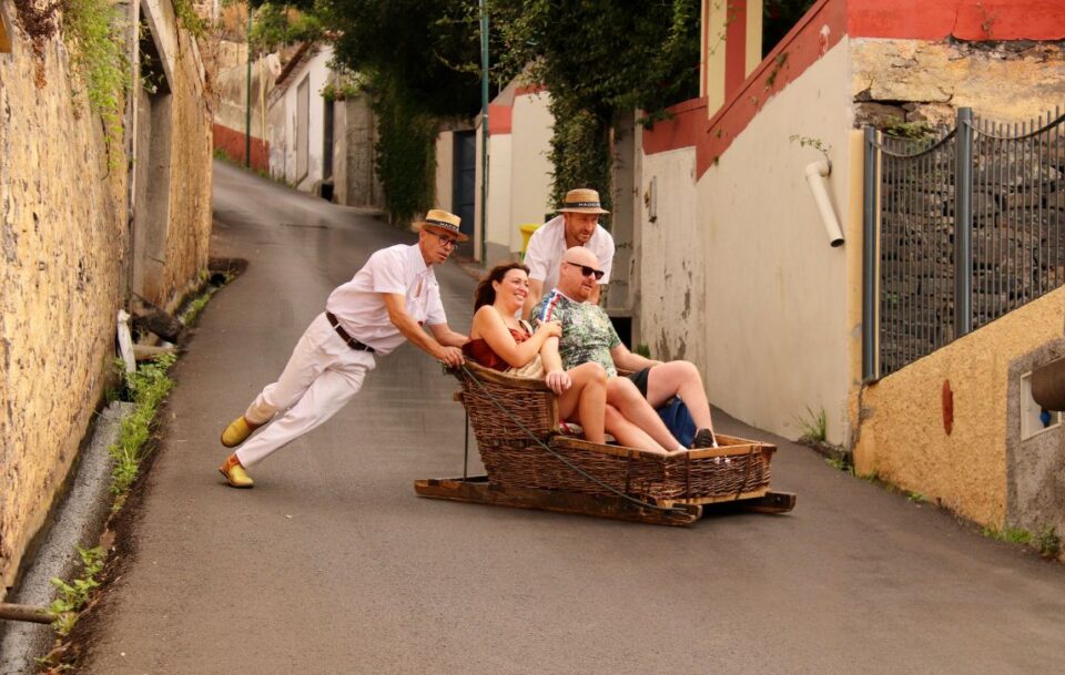 Wicker sled with tourists gliding down a steep street in Funchal on Madeira Island guided by two locals using their boots to steer and brake.